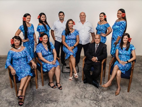 A group of ten people, wearing coordinated blue and white outfits, pose indoors for a formal photo. Four people are seated in chairs, whilst six stand behind them, all smiling at the camera.