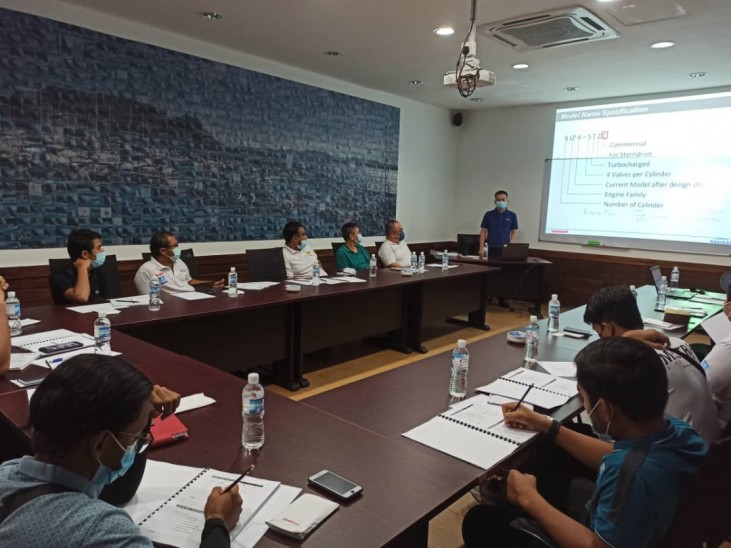 A group of people wearing masks sit around a conference table, listening to a presenter standing near a projection screen displaying technical information. Bottled water and documents are on the tables.