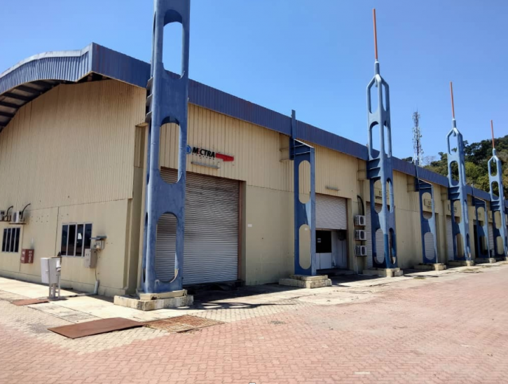 A large industrial warehouse with beige walls, blue structural supports, and closed grey roller shutters. The building stands on a paved area under a clear blue sky.