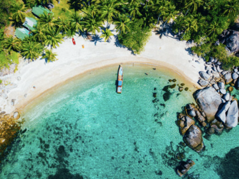 Aerial view of a tropical beach with turquoise water, a boat anchored near the shore, white sand, lush green palm trees, and large rocks along the coast.