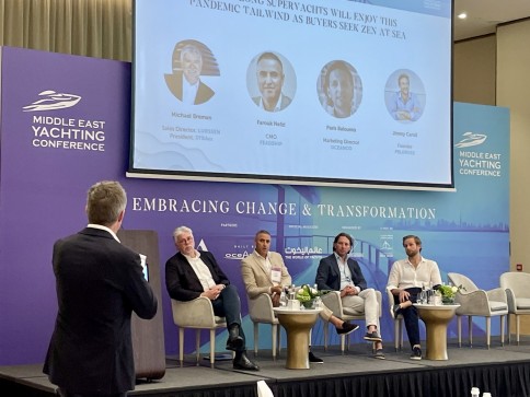 A man stands and speaks to a seated panel of four men at the Middle East Yachting Conference. A large screen behind them shows speaker names, photos, and the session title. The event backdrop features conference branding.