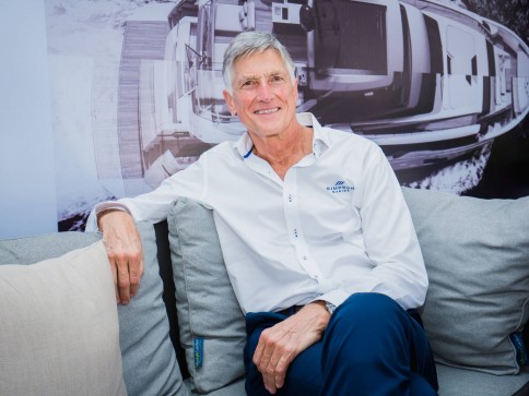 An older man with grey hair, wearing a white button-up shirt and blue trousers, sits on a light grey sofa and smiles. A black-and-white image of a yacht is displayed on the wall behind him.