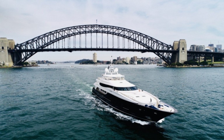 A large white yacht sails on the water with the Sydney Harbour Bridge in the background and city buildings visible on both sides of the bridge under a cloudy sky.