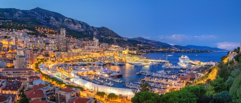 A panoramic view of Monaco at dusk, showing a brightly lit harbour filled with yachts, surrounding city buildings, hills in the background, and the sea stretching into the distance under a clear evening sky.