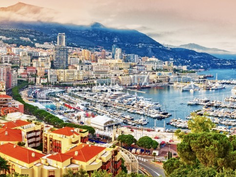 A scenic view of Monaco’s harbour with numerous yachts moored, colourful buildings along the waterfront, and mountains in the background partially covered by clouds.