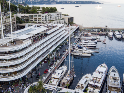Aerial view of a modern marina with large yachts moored next to a multi-storey building, bustling with people, near a coastline and a calm blue sea. Hills and more boats are visible in the background.