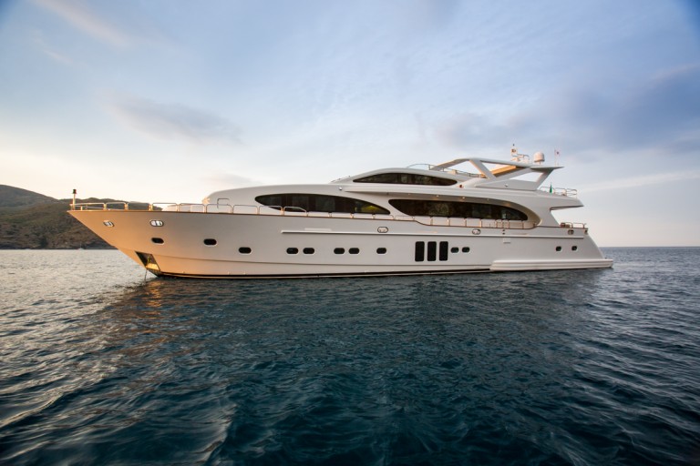 A large white luxury yacht is moored on calm blue water at sunset, with a partly cloudy sky and a distant shoreline visible in the background.