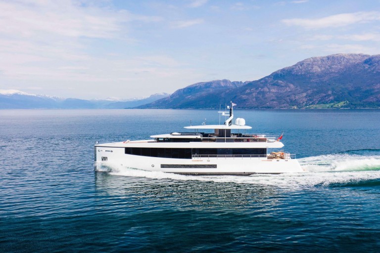 A modern white yacht cruises on calm blue water, leaving a trail behind, with green mountains and a partly cloudy sky in the background.
