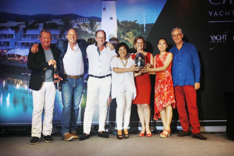 Seven adults stand on a stage in front of a backdrop with a marina scene. Four women and three men pose smiling, with two women holding a trophy together. Everyone is dressed in smart casual or semi-formal attire.