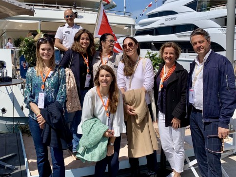 A group of seven people, six women and one man, smile and pose together on a dock with yachts in the background. Most are wearing jackets and orange lanyards with name badges. The weather appears sunny and pleasant.