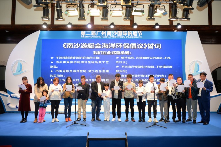 A group of adults and children stand on a stage in front of a blue backdrop with Chinese text, holding papers and reading aloud at the Guangzhou Nansha International Sailing Festival.