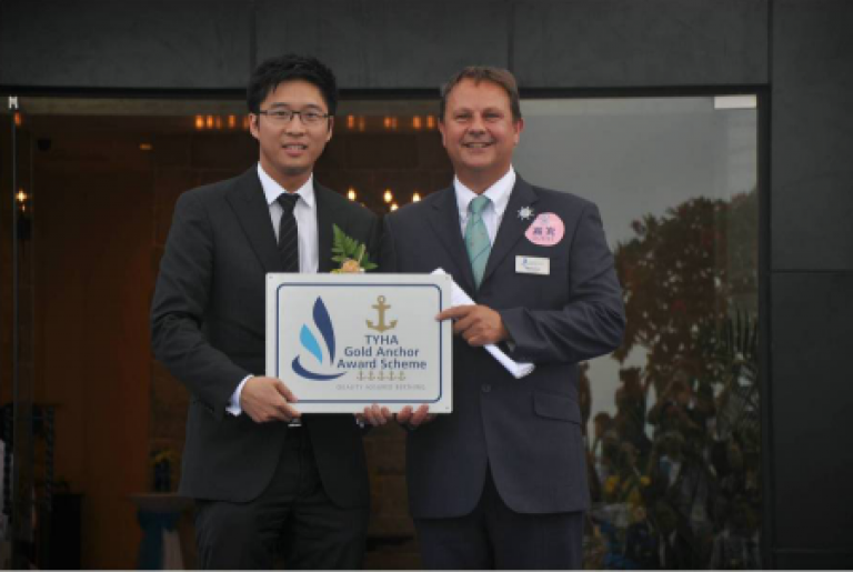 Two men in suits stand side by side, smiling and holding a plaque that reads “TYHA Gold Anchor Award Scheme.” One man wears glasses and a buttonhole; the other wears a name badge and a round pink badge.