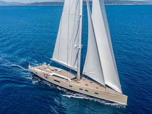 A large, modern sailboat with white sails glides across calm blue ocean water under a clear sky, with distant land visible on the horizon.
