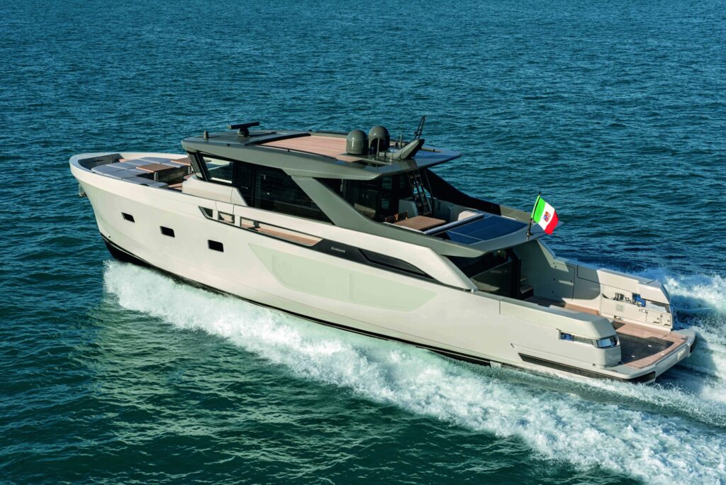 A modern motor yacht cruises on blue water, leaving a white wake behind. The vessel has a sleek design, several decks, and an Italian flag flying at the stern.