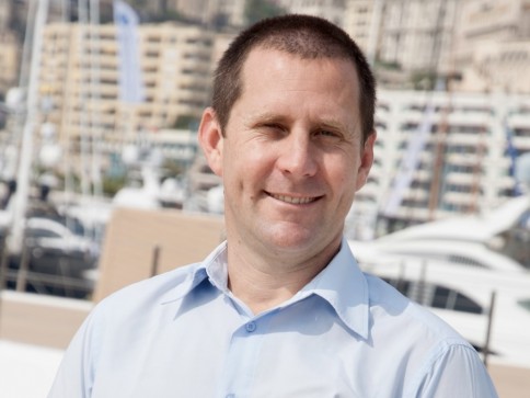 A man in a light blue shirt smiles outdoors with a marina and yachts in the background, and buildings on a hillside.