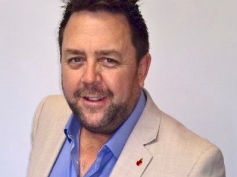 A man with short dark hair and a beard, wearing a light beige jacket over a blue shirt, smiles at the camera against a plain light background.