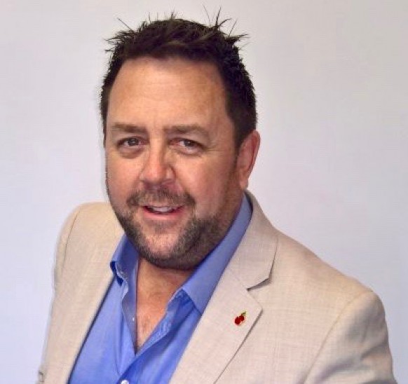 A man with short dark hair and a trimmed beard, wearing a light beige jacket and a blue shirt, smiles at the camera against a plain light background.