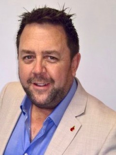 A man with short, dark hair and a trimmed beard is smiling. He is wearing a light beige blazer over a blue shirt, and there is a small red pin on his lapel. The background is plain and light coloured.