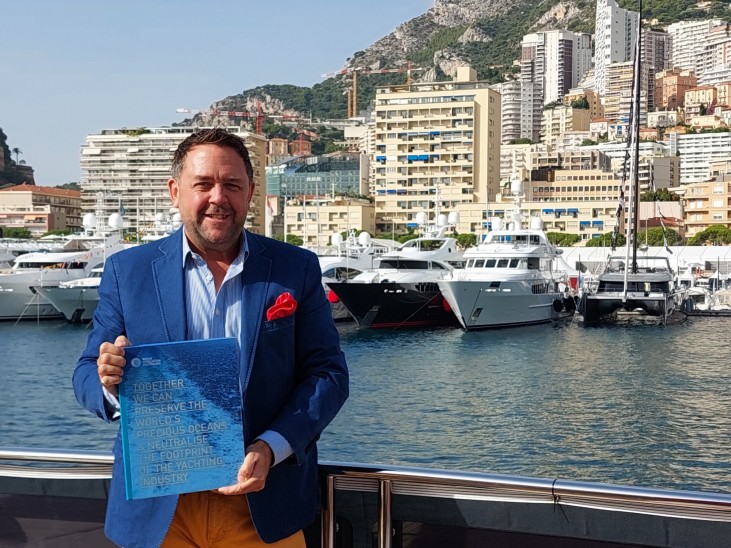 A man in a blue blazer and yellow trousers holds a blue booklet while standing by a marina with yachts and city buildings in the background, set against a hillside.