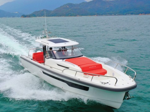 A sleek white motorboat with red seating speeds across a calm, green-blue sea, leaving a white wake behind. Green mountains are visible in the background under a cloudy sky.