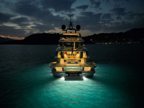 A luxury yacht is illuminated by underwater lights, casting a blue glow on the surrounding water at dusk. The sky is partly cloudy, and distant hills and city lights are visible in the background.