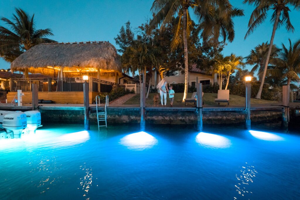 A waterfront scene at dusk with glowing blue underwater lights along a quay, palm trees, a thatched-roof structure, people walking, and loungers near the water’s edge.