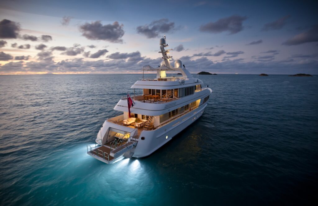 A luxury yacht illuminated with soft lights floats on calm ocean waters at sunset, with a dramatic sky and small islands visible in the distance.