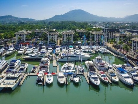 A marina filled with numerous yachts and boats moored in green-blue water, with modern buildings and palm trees in the background, and hills rising under a clear blue sky.