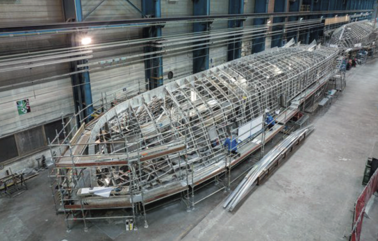 A large metal ship frame under construction inside a spacious industrial building, surrounded by scaffolding, construction materials, and equipment.