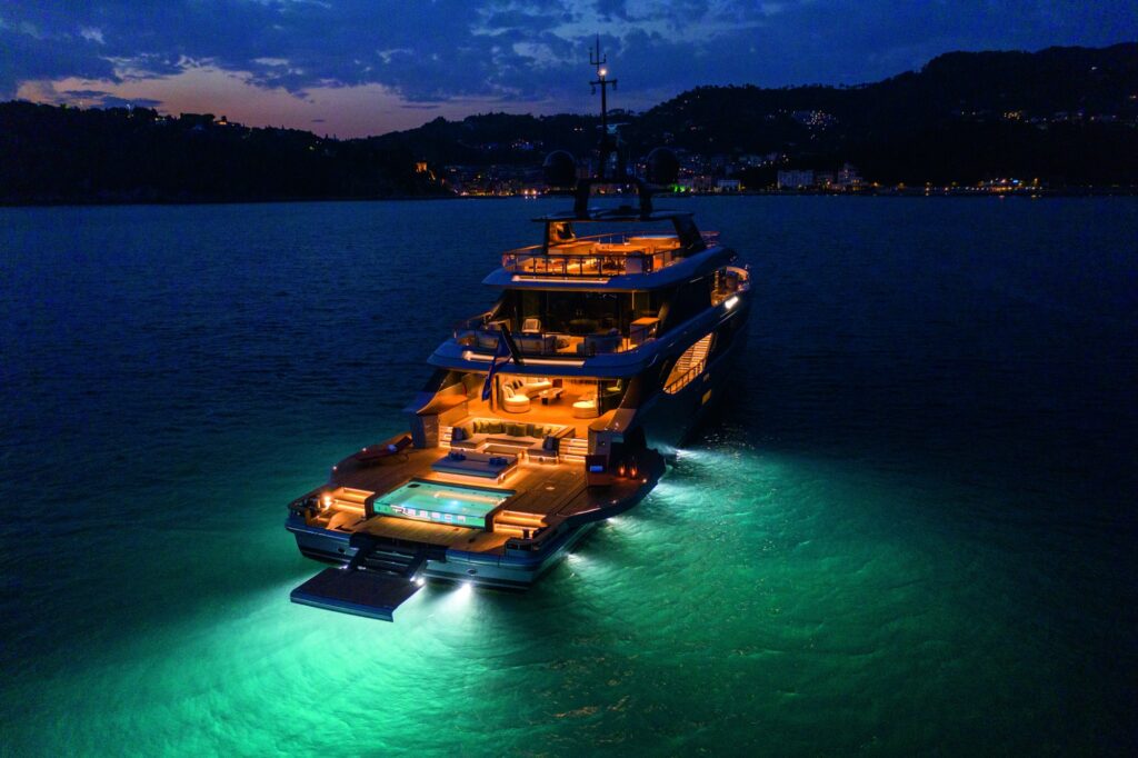 A luxurious yacht with glowing underwater lights and illuminated decks floats on calm water at dusk, with distant hills and scattered lights visible along the shoreline.