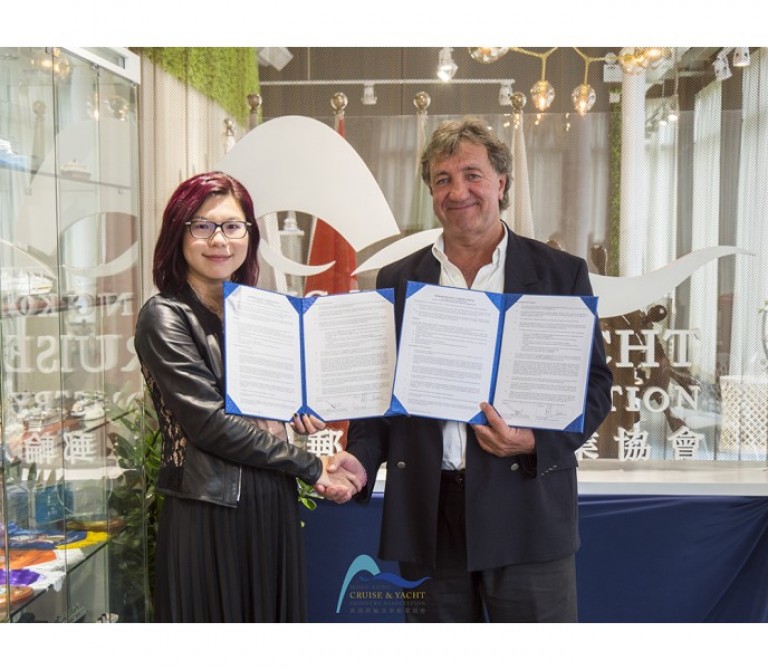 A woman and a man stand side by side, smiling and shaking hands whilst holding an open document together. They are indoors, in front of a display case and a logo backdrop.