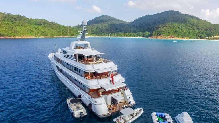 A large white luxury yacht is anchored near a lush, green island in clear blue water. Smaller boats and jet skis are moored alongside the yacht, with forested hills and a sandy beach visible in the background.