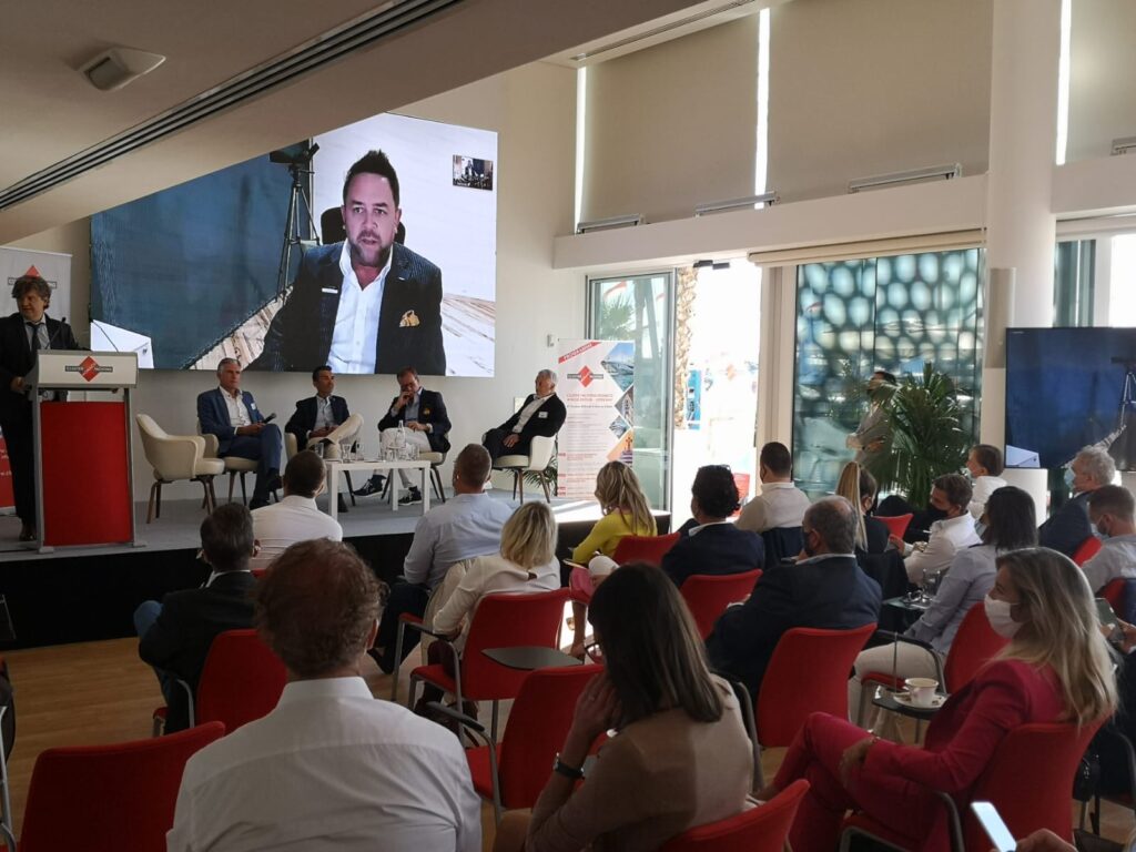 A business conference with attendees seated facing a stage, where speakers sit on a panel. One speaker appears virtually on a large screen. Most attendees wear business attire; some wear masks. Daylight streams through tall windows.