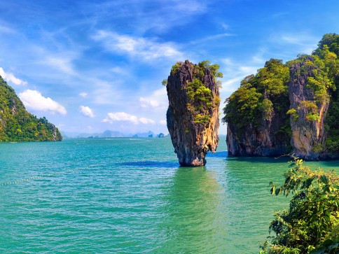 A tall, narrow limestone island rises from turquoise water, surrounded by lush green cliffs under a bright blue sky with scattered clouds.