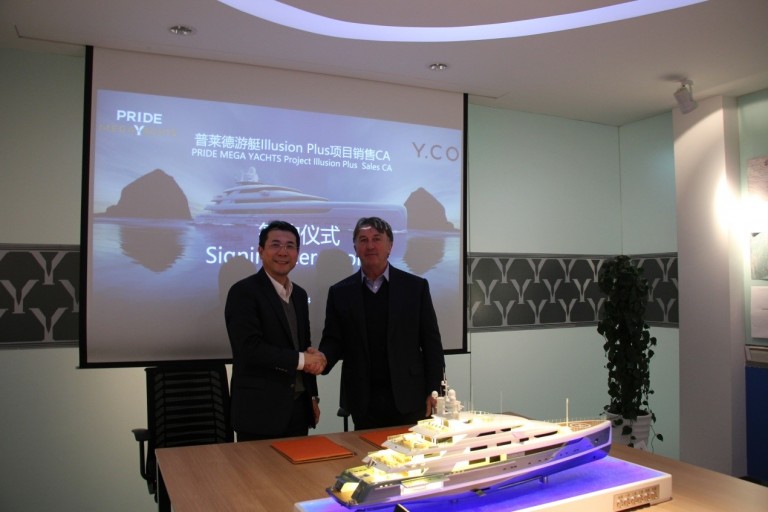 Two men in suits shake hands in an office. Behind them, a digital screen displays a yacht and the words PRIDE MEGA YACHTS Project Illusion Plus Sales CA. A detailed yacht model sits on the table in front of them.