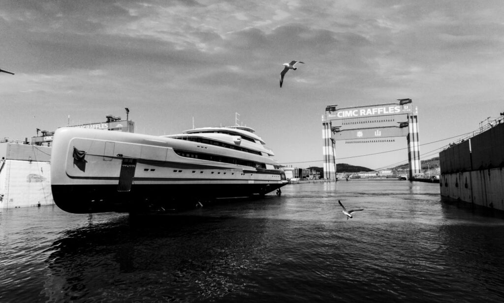 A large yacht is moored at a shipyard with a CIMC Raffles gantry crane overhead. Two seagulls fly above the water under a partly cloudy sky. The image is in black and white.