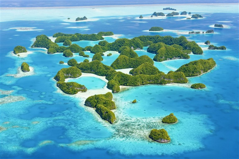 Aerial view of small, lush green islands scattered across clear turquoise blue water, surrounded by coral reefs and shallow sandy areas, under a bright sky.