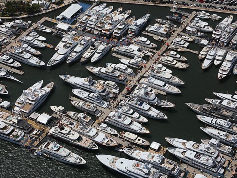 Aerial view of a crowded marina with dozens of large, white yachts moored side by side in multiple rows along several piers, with people walking along the jetties.