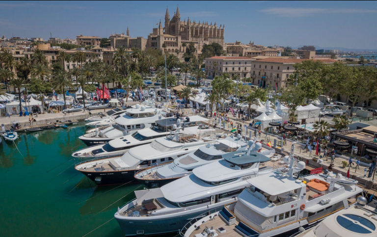 A marina filled with luxury yachts, with a bustling crowd on the promenade. Historic buildings and a large cathedral with gothic features are visible in the background under a blue sky.