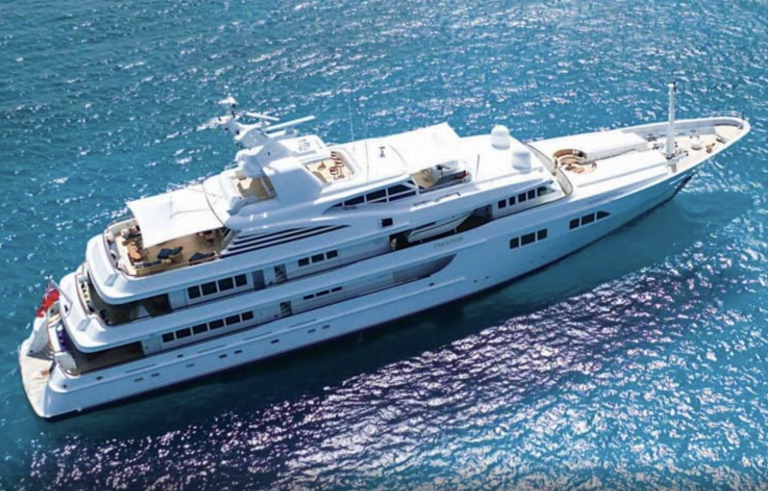A large, luxurious white yacht with multiple decks floats on clear blue ocean water, viewed from above. The yacht features lounging areas and sun decks, with the sun reflecting off the sea.
