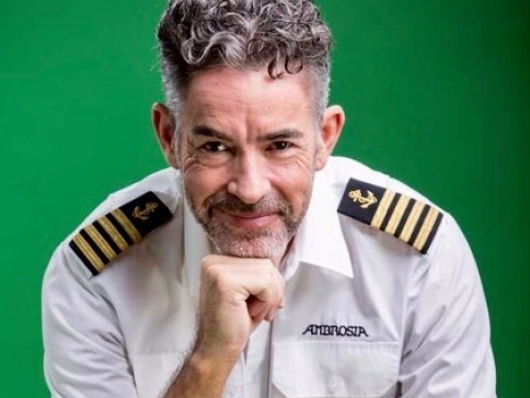 A middle-aged man with curly grey hair and a beard, wearing a white captain's uniform with shoulder epaulettes, smiles whilst resting his chin on his hand against a plain green background.