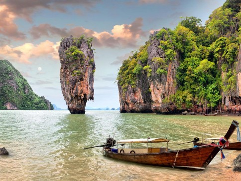 A wooden long-tail boat is moored in shallow water near a lush, green island with a tall limestone rock formation rising from the sea under a cloudy sky.