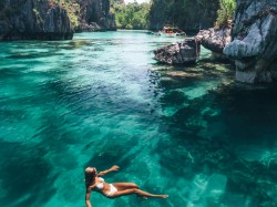 A woman in a white bikini floats on clear turquoise water surrounded by dramatic rocky cliffs and lush greenery, with a small boat in the background.