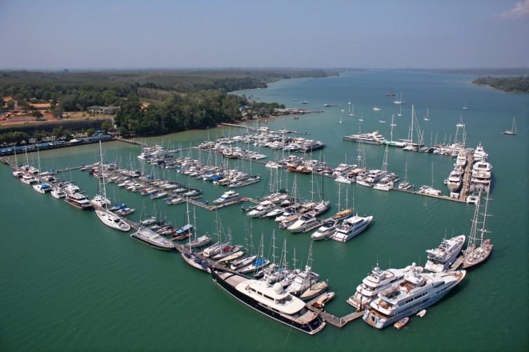 Aerial view of a large marina filled with numerous yachts and boats moored in turquoise water, surrounded by green trees and land, with a wide river or bay stretching into the distance under a hazy sky.