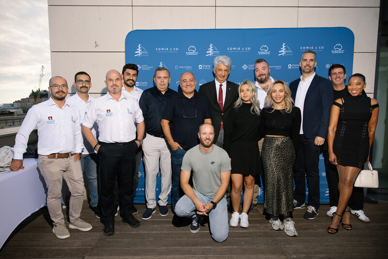 A group of thirteen people pose and smile together on a wooden decking in front of a blue Cowie & Co event backdrop. They are dressed in business casual to formal attire, some standing and some kneeling.