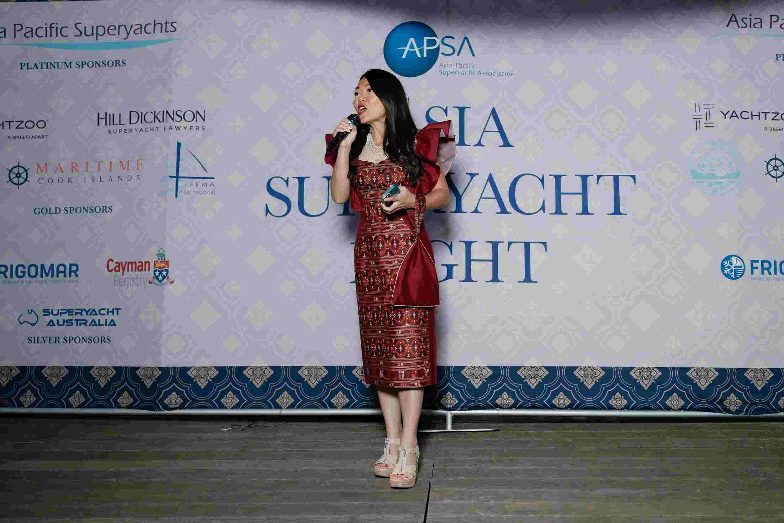 A woman in a red patterned dress holds a microphone and a matching handbag whilst standing on stage in front of a backdrop that reads Asia Superyacht Night with various sponsor logos.