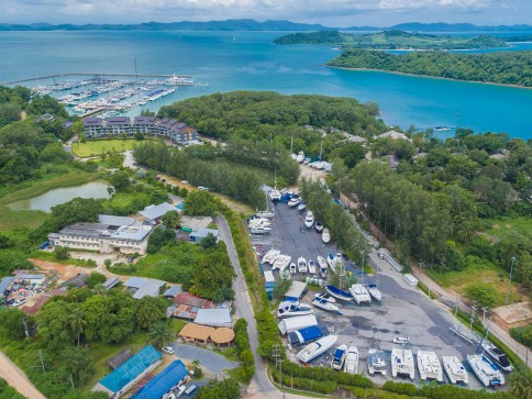 Aerial view of a marina with many moored boats, surrounded by green trees and buildings, adjacent to a turquoise bay and distant forested islands under a partly cloudy sky.
