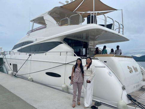 Two women stand smiling on a quay in front of a large white yacht. Several people are visible on the yacht’s deck. The sky is overcast and the water is calm in the background.