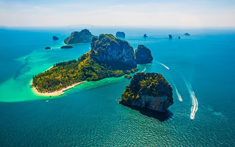 Aerial view of rocky islands surrounded by turquoise sea water, with lush greenery, sandy beaches, and speedboats creating white trails on the water under a clear blue sky.
