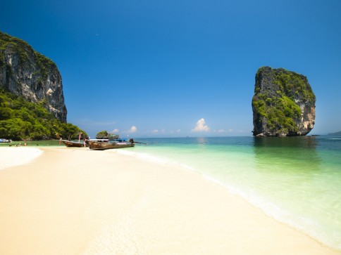 A tropical beach with white sand, clear turquoise water, longtail boats near the shore, and large limestone cliffs covered in greenery under a bright blue sky.